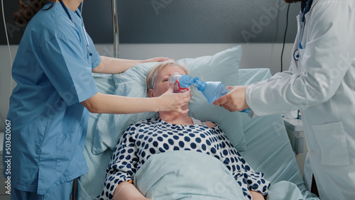 Doctor and nurse using oxygen tube for hyperventilating woman in bed. Medical specialists with tool and heart rate monitor for reanimation and respiratory problem of senior patient.