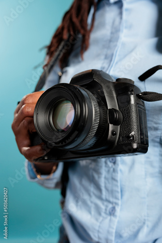 Close up of young adult photograhpy entusiast holding a modern camera device while standing on blue background. Professional photographer with DSLR enjoying leisure time. Studio shot