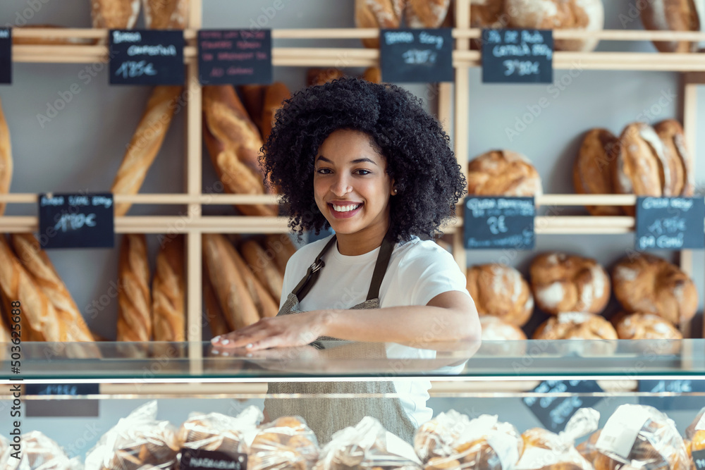 Beautiful afro younger owner woman selling fresh pastry and loaves un bread section and smiling at pastry shop.
