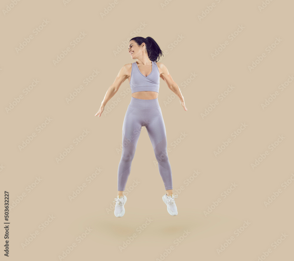 Dynamic movement. Healthy sport woman with fit body doing fitness jumps isolated on beige background. Smiling young woman in sportswear doing jumping exercise. Workout sport concept. Full length. Dynamic movement. Healthy sport woman with fit body doing fitness jumps isolated on beige background. Smiling young woman in sportswear doing jumping exercise. Workout sport concept. Full length.