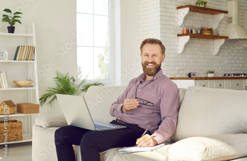 Happy man working on his laptop computer at home. Cheerful handsome bearded businessman or distant, remote student sitting on a comfortable sofa, using his notebook PC and taking notes in a notepad