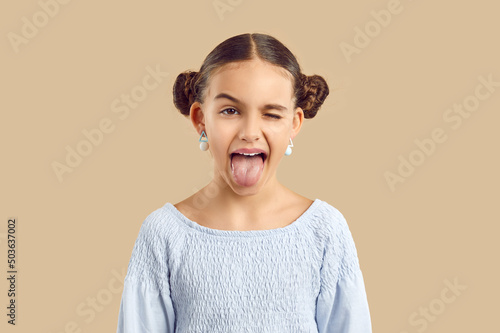 Happy child sticking tongue out. Studio shot of a cheerful naughty kid making a funny grimace. Pretty little girl with adorable space hair buns and modern earrings shows her tongue and winks her eye