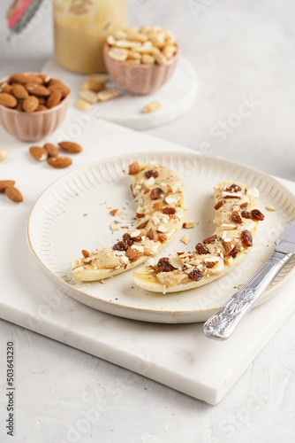 Two banana halves ready with peanut butter topping, raisins, almonds and chia seeds on white plate on marble board