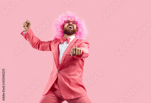 Cheerful crazy man in pink wig making funny dance moves isolated on pink background. Eccentric man in pink formal suit rejoices, has fun and dances in gangnam style. Banner. photo
