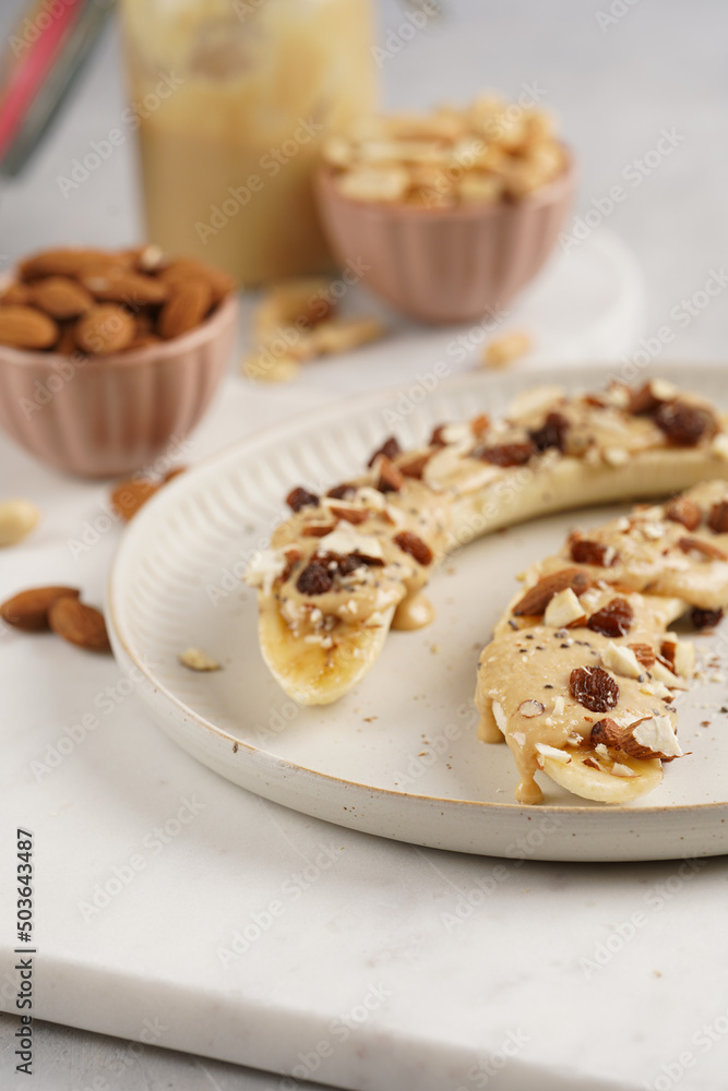 Two banana halves ready with peanut butter topping, raisins, almonds and chia seeds on white plate on marble board Two banana halves ready with peanut butter topping, raisins, almonds and chia seeds on white plate on marble board