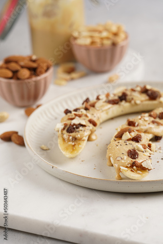 Two banana halves ready with peanut butter topping, raisins, almonds and chia seeds on white plate on marble board