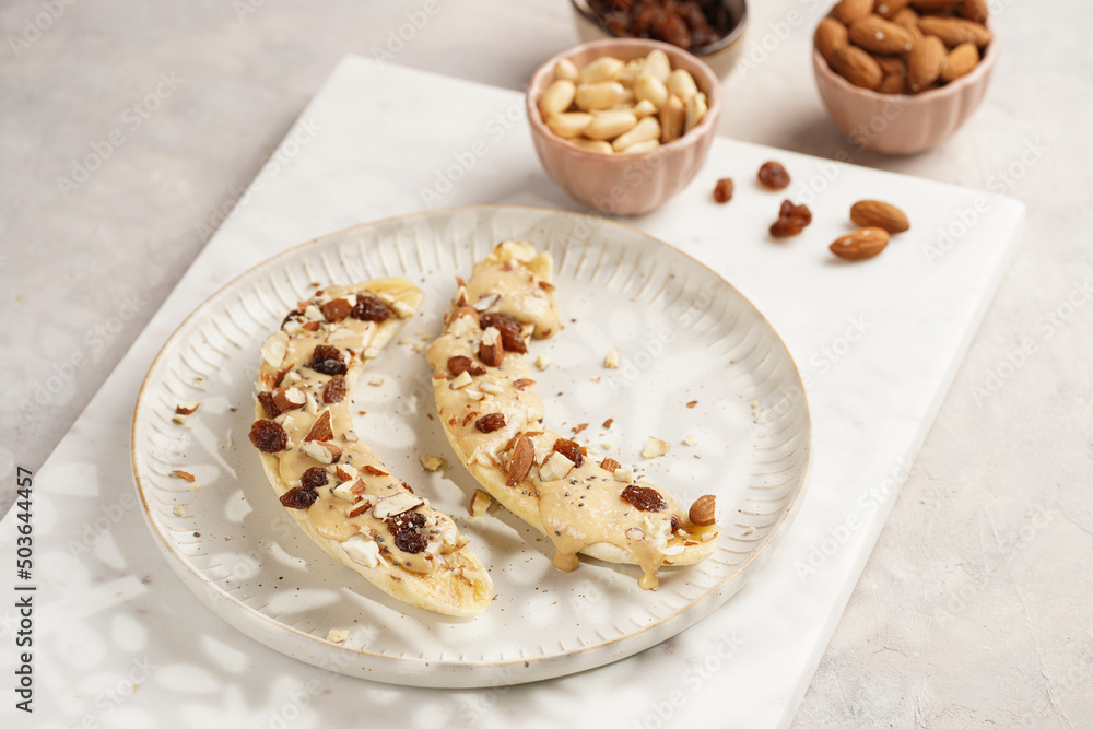 Two banana halves ready with peanut butter topping, raisins, almonds and chia seeds on white plate on marble board, direct creative sunlight shadow