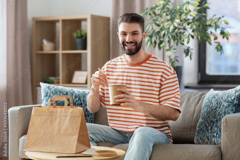 consumption, delivery and people concept - smiling man with chopsticks eating takeaway food at home