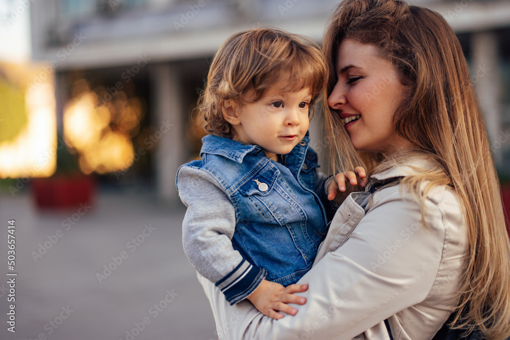 Fototapeta premium Side profile of a beautiful young mother, holding her male toddler, outdoor.