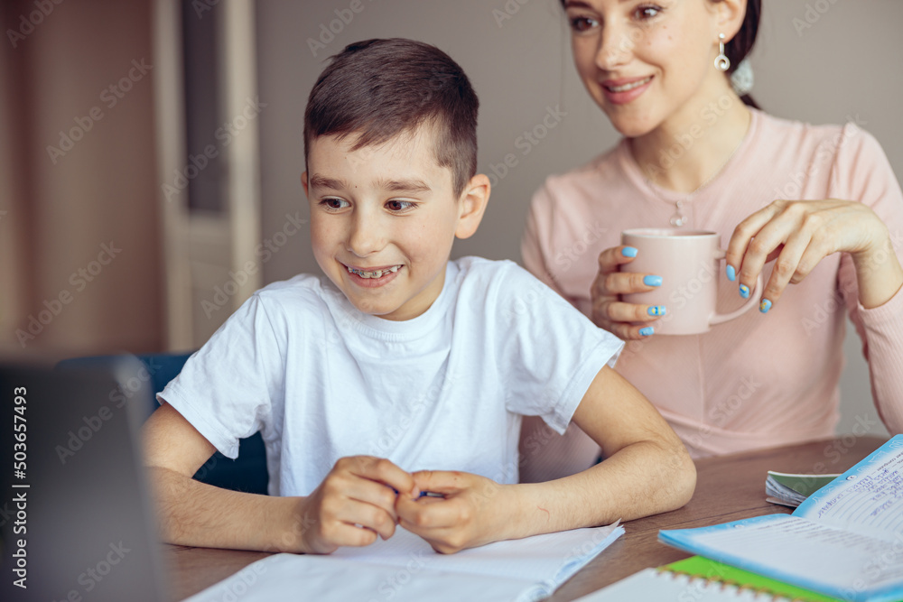 Little happy boy with braces smiling and looking at laptop. Mom drinking coffee. Little happy boy with braces smiling and looking at laptop. Mom drinking coffee.