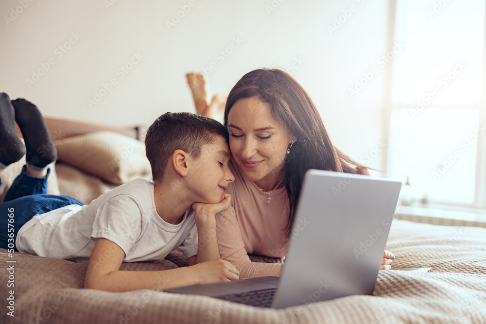 Happy Caucasian smiled mother and cute son cuddling while lying with laptop on bed. Happy Caucasian smiled mother and cute son cuddling while lying with laptop on bed.