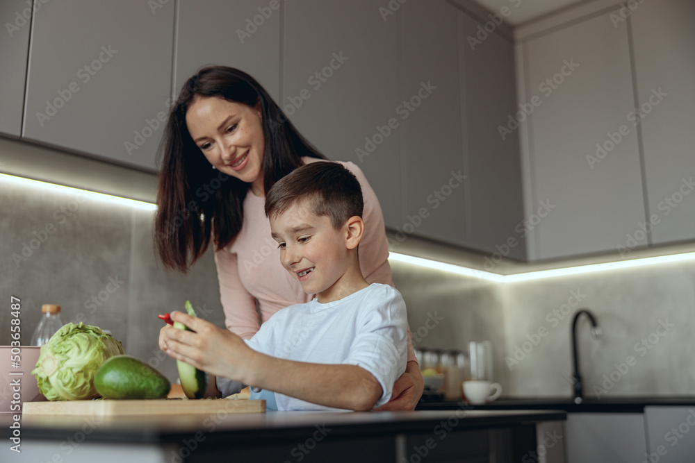 Cheerful young Caucasian mother with small son peeling vegetables and cooking.