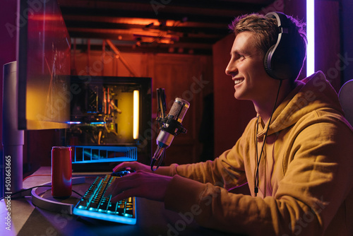  Excited caucasian gamer streamer with headphones playing computer pc video at his living room, recording live stream using microphone, drinking energy drink. Cyber sport and social media concept photo