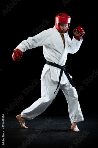 Male taekwondo fighter in white dobok, helmet and gloves training isolated over dark background. Concept of sport, workout, competition, ad