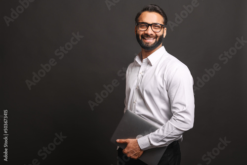 Successful Indian businessman in formal wear and glasses carrying laptop computer standing isolated on black background, portrait of proud hispanic male entrepreneur