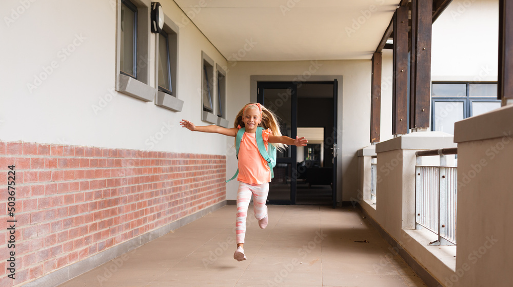 Cheerful caucasian elementary schoolgirl with arms outstretched running in school corridor