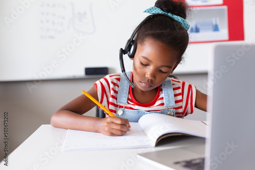African american elementary schoolgirl wearing headphone while writing on book at desk with laptop