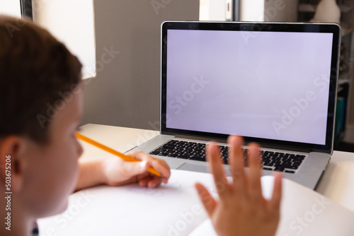 Caucasian elementary schoolboy waving hand on laptop while studying over video conference