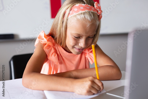 Smiling caucasian elementary schoolgirl writing on book while studying with laptop at desk in class