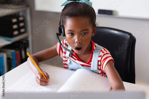 African american elementary schoolgirl with mouth open looking at laptop while studying at desk