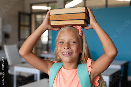 Portrait of playful caucasian elementary schoolgirl with stacked books on head standing in class
