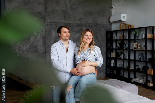 Photo of joyful young man and pregnant woman couple wait baby touch belly sit sofa indoors living room house