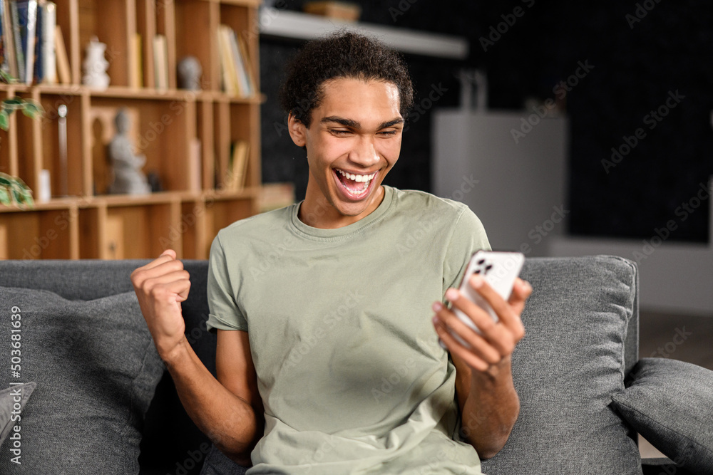 Excited multiracial man sitting on the sofa looking at the phone and smiling, surprised emotional lucky man, rejoicing at good message, news, won the lottery Excited multiracial man sitting on the sofa looking at the phone and smiling, surprised emotional lucky man, rejoicing at good message, news, won the lottery