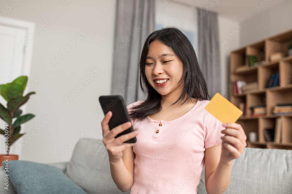 Cheerful korean lady with mobile phone and credit card purchasing goods or services on web sitting on sofa at home