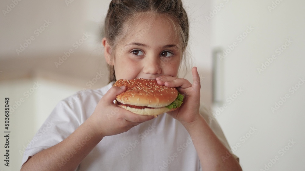 little girl eating a hamburger. unhealthy fast food proper nutrition concept. child greedily with pleasure lifestyle bites a big burger in the kitchen at home. kid eats fast food close-up