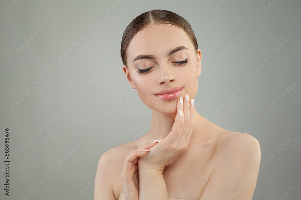 Young woman with natural makeup and healthy skin. Beautiful spa model touching hydrated facial skin on gray background. Skin care and cosmetology concept