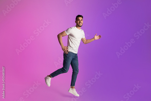 Full length of happy young Arab man in t-shirt and jeans running in neon light, copy space