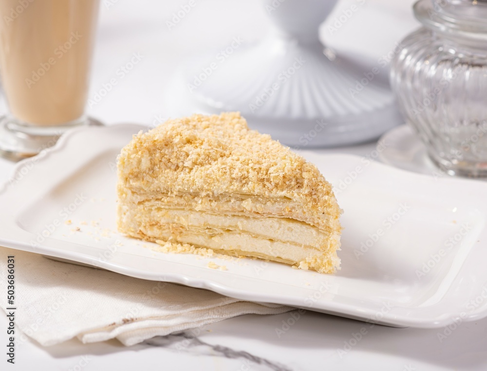 A piece of Napoleon cake on a light marble table among white dishes A piece of Napoleon cake on a light marble table among white dishes