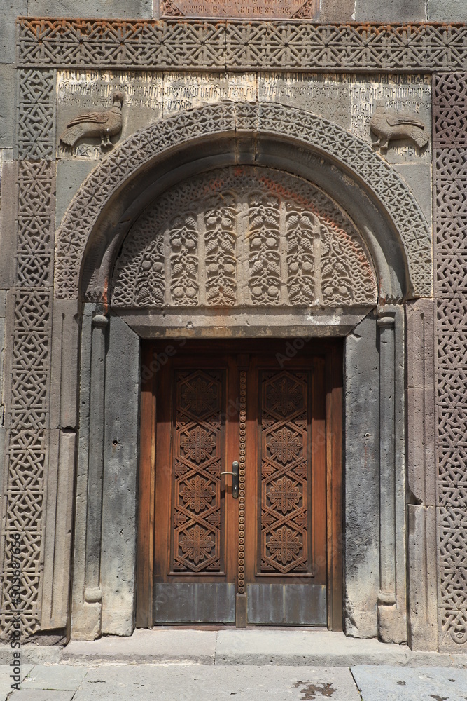 Facade and entrance of the main temple in the ancient monastery of Geghard in the mountains of Armenia, built into the rock. The wooden door is decorated with carvings and framed with an arched vault.