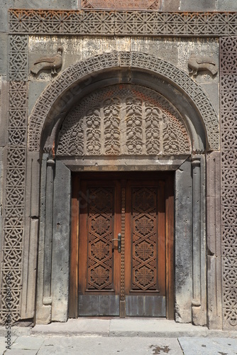 Facade and entrance of the main temple in the ancient monastery of Geghard in the mountains of Armenia, built into the rock. The wooden door is decorated with carvings and framed with an arched vault.