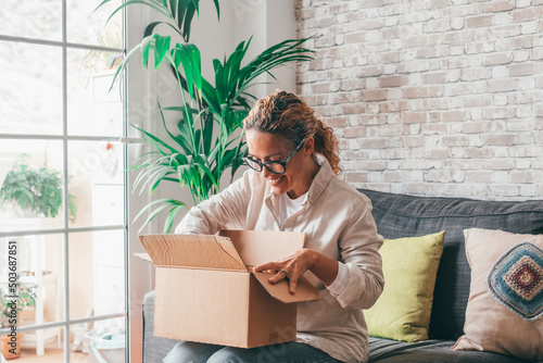 Overjoyed woman seated on sofa hold on lap small cardboard box open parcel client feels satisfied bought goods in internet, happy addressee received package from friend, quick delivery service concept photo