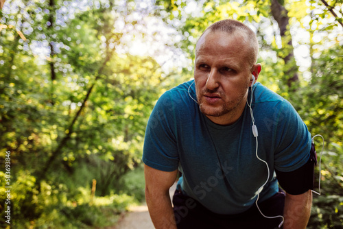 Caucasian man resting after run at park with copy space while listening to music. Handsome runner with earphones in his ears, listening music, outside in sunny autumn nature, resting, breathing out. © Jelena Stanojkovic