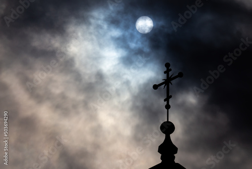 Orthodox cross on top of the church against the dramatic sky. Photo taken against the sun behind the clouds.