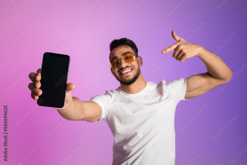 Smiling young Arab man in sunglasses pointing at smartphone with empty screen in neon light, selective focus. Mockup Smiling young Arab man in sunglasses pointing at smartphone with empty screen in neon light, selective focus. Mockup