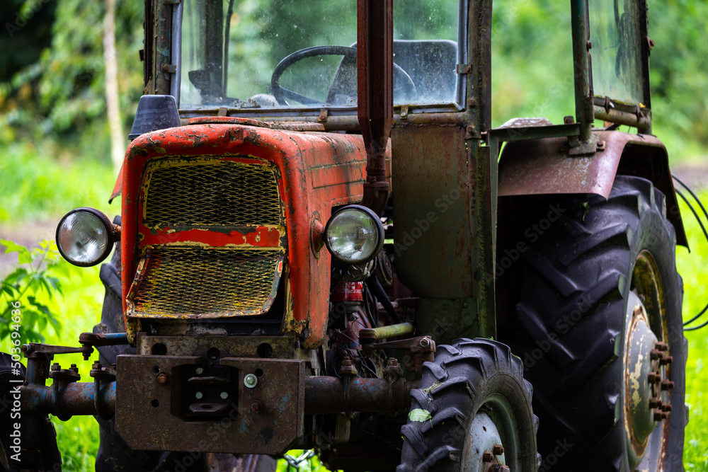 An old red tractor standing in the shade among the trees. The photo was taken on a cloudy day with good lighting conditions.