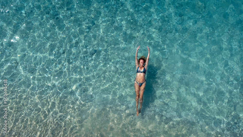 Aerial view of beautiful happy woman in swimsuit laying in the shallow sea water enjoying beach and soft turquoise ocean wave. Tropical sea in summer season on Egremni beach on Lefkada island.