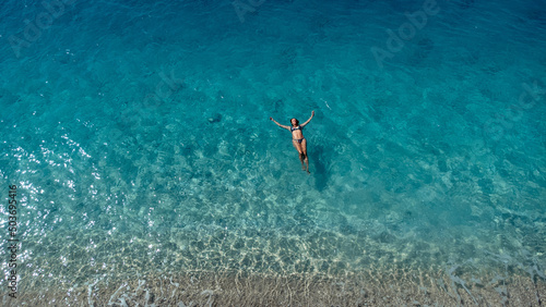 Aerial view of beautiful happy woman in swimsuit laying in the shallow sea water enjoying beach and soft turquoise ocean wave. Tropical sea in summer season on Egremni beach on Lefkada island. © MexChriss
