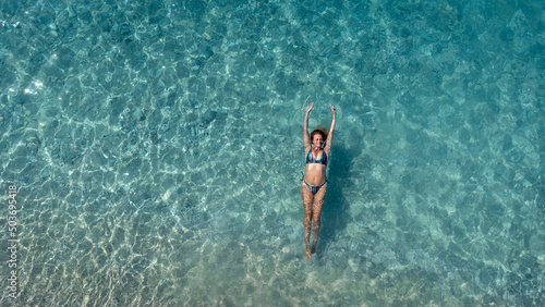 Aerial view of beautiful happy woman in swimsuit laying in the shallow sea water enjoying beach and soft turquoise ocean wave. Tropical sea in summer season on Egremni beach on Lefkada island.