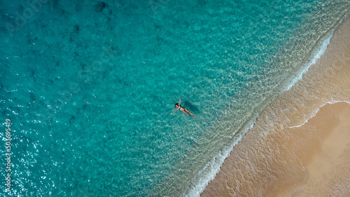Aerial view of beautiful happy woman in swimsuit laying in the shallow sea water, enjoying sandy beach and soft turquoise ocean wave. Tropical sea in summer season on Egremni beach on Lefkada island. © MexChriss