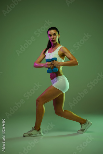 Portrait of young sportive girl doing squats with dumbbell isolated over green studio background in neon light