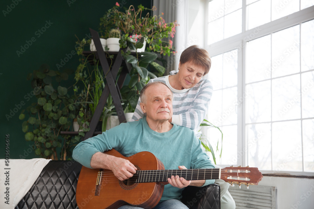 Fototapeta premium elderly couple spend time together. healthy lifestyle. emotional health. rest from gadgets. an elderly man plays guitar for his wife sitting on the couch at home. Slow life. Enjoying the little things