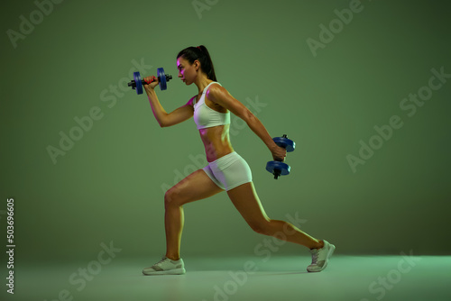 Side view portrait of young sportive girl training, doing leg exercises with dumbbell isolated over green background in neon