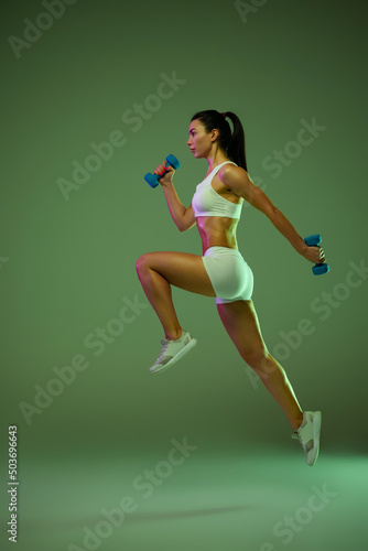 Full-length portrait of young muscular girl training, jumping with dumbbells isolated over green background in neon light