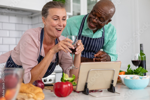 Happy senior multiracial couple with wine watching online recipe and preparing food at home