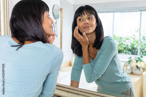 Caucasian young woman looking in mirror while applying moisturizer on face at home