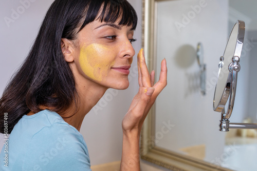 Caucasian young woman looking at mirror and applying facial mask in bathroom